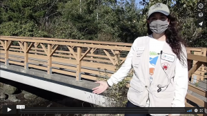 Naturalist Carolina stands in front of the wooden railing of a bridge, with green trees in the background, and a stream below. She wears a vest, a ballcap, and a facemask, and has long brown hair.
