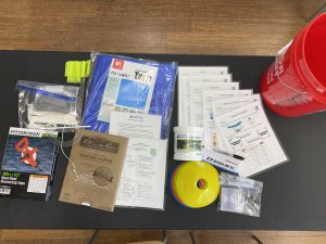 Display of Teacher PD kit of materials including a water quality testing kit and instructions, rite in the rain paper, cones and flagging, tarp, clinometer kit, measuring tape, and red bucket to store everything.